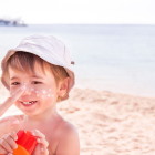 Urlaubsziele für Reisen mit Kindern / Mutter cremt Kind am Strand mit Sonnencreme ein – Sommerurlaub mit Kindern