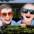 Autoreise mit Kindern / Zwei Kinder lachen mit Sonnenbrillen aus dem geöffneten Autofenster während einer Autofahrt