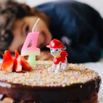 4. Geburtstag, 5. Lebensjahr / Geburtstagstorte mit einer Kerze in Form der Zahl 4, dahinter schaut ein Kind lächelnd auf den Kuchen