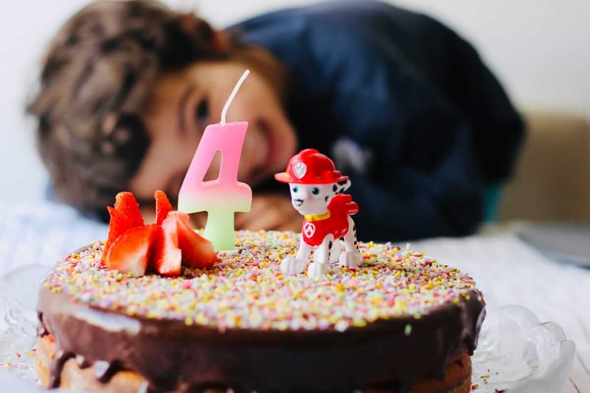 4. Geburtstag, 5. Lebensjahr / Geburtstagstorte mit einer Kerze in Form der Zahl 4, dahinter schaut ein Kind lächelnd auf den Kuchen