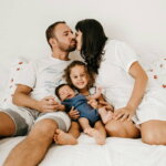 Eine glückliche Familie liegt zusammen auf einem Bett. Die Mutter gibt dem Vater einen Kuss, während das kleine Kind zwischen ihnen sitzt und lächelt. Ein Neugeborenes liegt entspannt auf dem Schoß des Vaters.