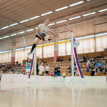 Hobby Horsing / Ein Mädchen springt mit einem Hobby Horse über eine Hürde in einer Halle. Zuschauer sind im Hintergrund zu sehen, während sie einen Moment in der Luft einfängt.