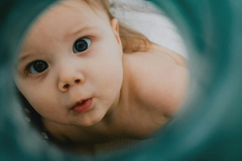 Top 10 2024 / Top Artikel (Nahaufnahme eines Babys mit großen, neugierigen Augen, das durch eine unscharfe, türkisfarbene Öffnung in die Kamera schaut.)