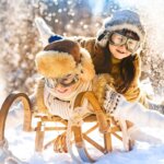 Pepco Kindermode (Zwei lachende Kinder in Winterkleidung mit Fellmützen und Schneebrillen rodeln fröhlich auf einem Holzschlitten durch den Schnee.)