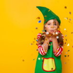 Karneval, Sicherheit / Ein fröhliches Mädchen im grünen Elfenkostüm bläst Konfetti in die Luft. Die bunten Sterne fliegen um sie herum vor einem leuchtend gelben Hintergrund.