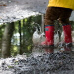 Frühling, spielen / Kind in einer gelben Regenjacke und roten Gummistiefeln springt in eine große Pfütze, wodurch das Wasser spritzt. Die Umgebung ist eine nasse, matschige Waldstraße.