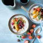 Brainfood (Zwei Schalen mit Joghurt und Haferflocken, garniert mit frischen Früchten wie Erdbeeren, Orangen und Passionsfrucht, auf einem blauen Holztisch, umgeben von Beeren, Sternanis und Löffeln.)