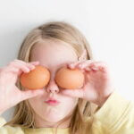 Ostern (Mädchen hält zwei braune Eier vor die Augen und zieht eine lustige Grimasse, vor hellem Hintergrund.)