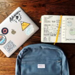 Lernmethoden (Laptop mit Aufklebern, aufgeschlagenes Notizbuch mit handschriftlichen Notizen und Diagrammen sowie ein blauer Rucksack auf einem Holztisch.)