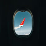 Flugreise (Blick aus dem Flugzeugfenster auf den Flügel einer Maschine der Fluggesellschaft Norwegian vor blauem Himmel mit Wolken.)