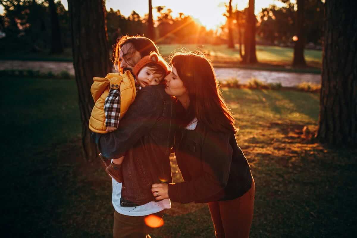 Kinderfotos / Eltern küssen ihr Kind im warmen Abendlicht im Park – liebevoller Familienmoment im Sonnenuntergang.