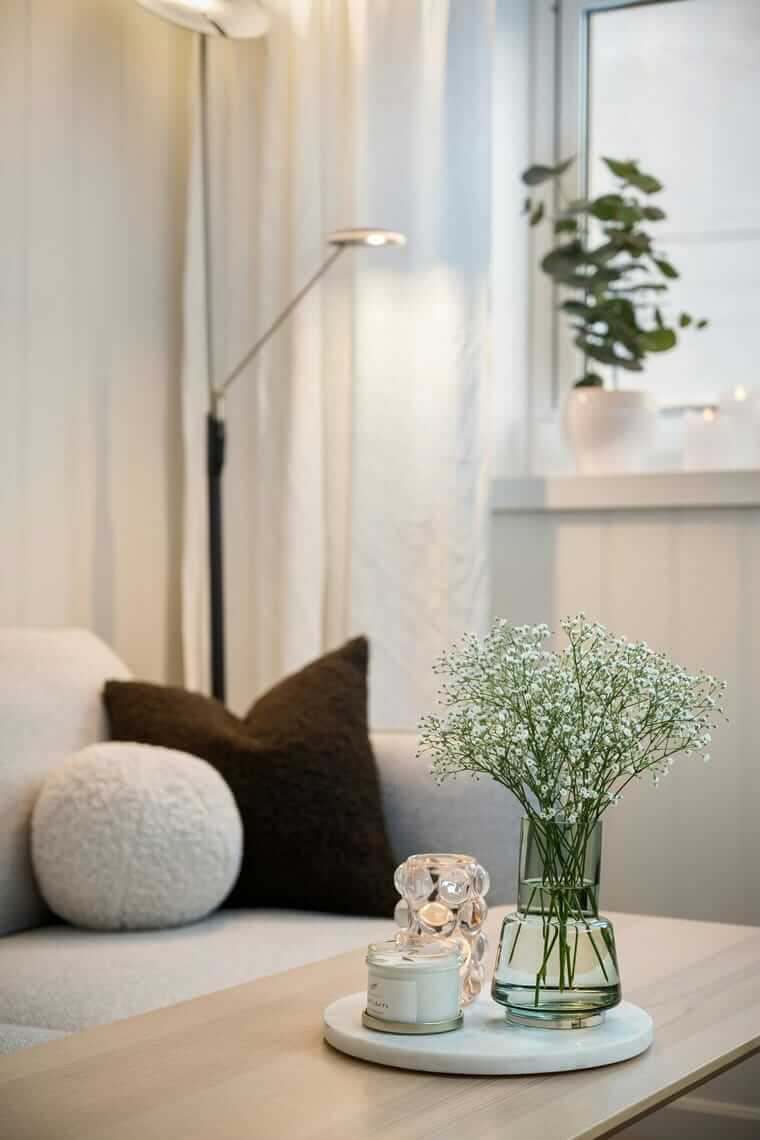 gemütlich zuhause / Helles, gemütliches Wohnzimmer mit weichem Sofa, beigefarbenen Kissen, Glasvase mit weißen Blüten und Kerze auf einem Tablett – ruhige, skandinavische Atmosphäre.