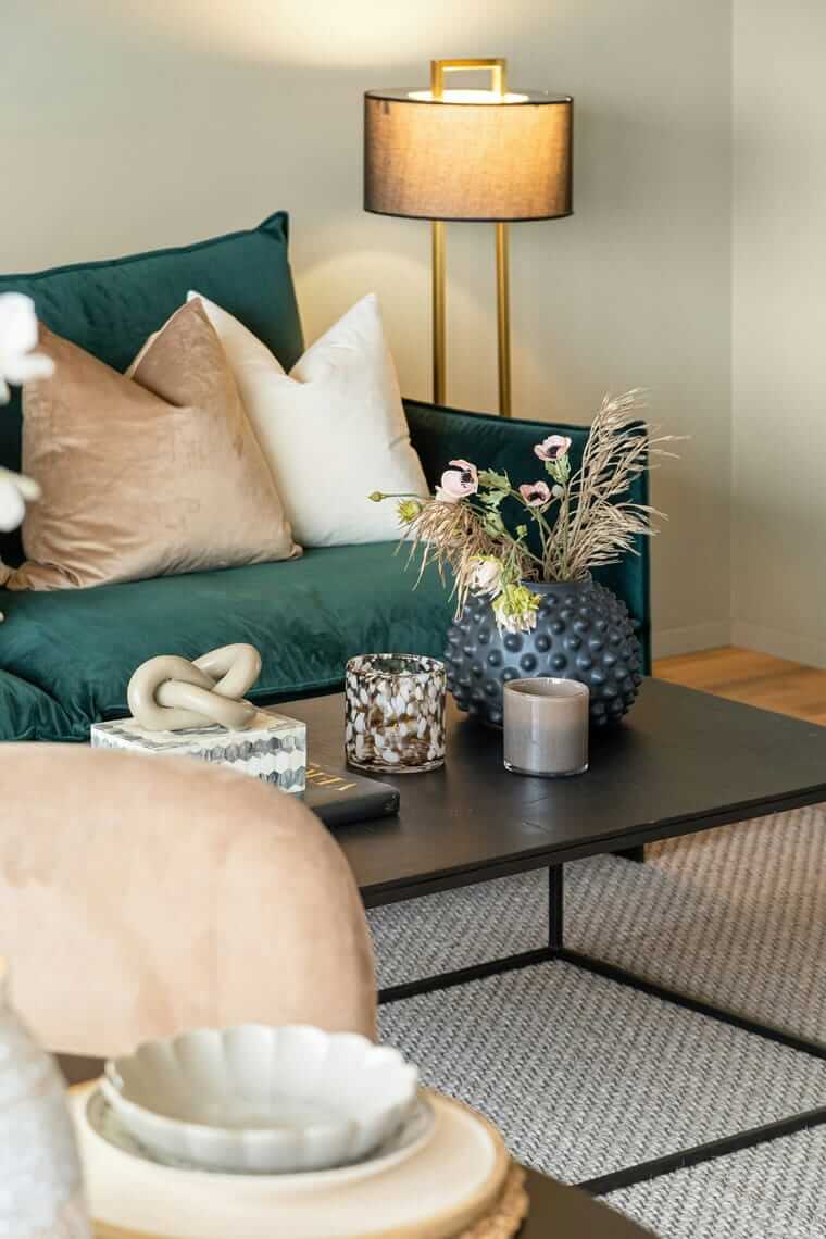 gemütlich zuhause / Detailaufnahme eines modernen Wohnzimmers mit dunkelgrünem Sofa, beigen Kissen, dekorativen Vasen auf dem Couchtisch und warmem Lampenlicht im Hintergrund.