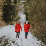 Zwei Kinder in roten Winterjacken laufen auf einem schneebedeckten Weg zwischen kahlen Sträuchern. / Schule, Schulweg, Winter