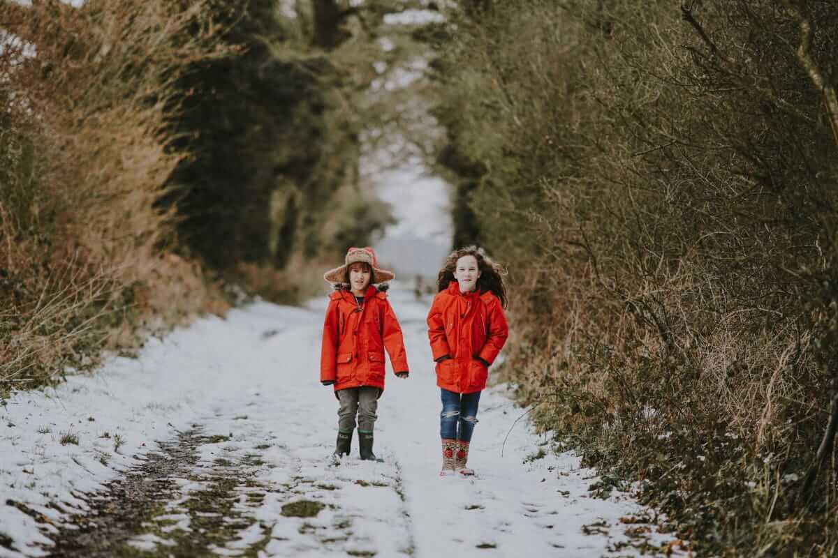 Zwei Kinder in roten Winterjacken laufen auf einem schneebedeckten Weg zwischen kahlen Sträuchern. / Schule, Schulweg, Winter