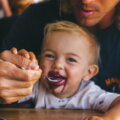 Geschmack (Kleines Kind probiert begeistert vom Löffel, während ein Elternteil hilft – gemeinsamer Genussmoment beim Essenlernen.)