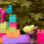 Kindergrundsicherung / Bunte Spielfiguren bauen farbige Bausteine zu unterschiedlich hohen Türmen – symbolisches Bild für Unterstützung und faire Startbedingungen für Kinder.