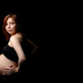 Schwanger mit 35 / Schwangere Frau Mitte 30 im Profil vor dunklem Hintergrund, selbstbewusster Blick in die Kamera, Hand am Bauch