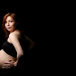 Schwanger mit 35 / Schwangere Frau Mitte 30 im Profil vor dunklem Hintergrund, selbstbewusster Blick in die Kamera, Hand am Bauch