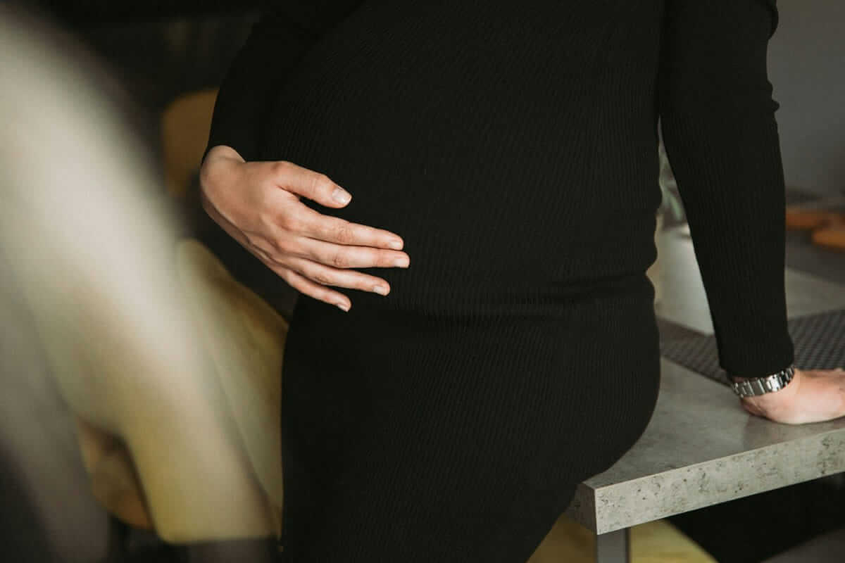 Schwanger mit 35, 38 oder 40: Was du wissen solltest Schwanger mit 35 / Schwangere Frau in schwarzem Strickkleid lehnt an Küchentheke, Hand ruht auf dem Bauch, Alltagsmoment