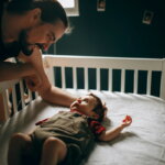 Reizhusten / Vater beugt sich nachts über das Gitterbett seines Babys