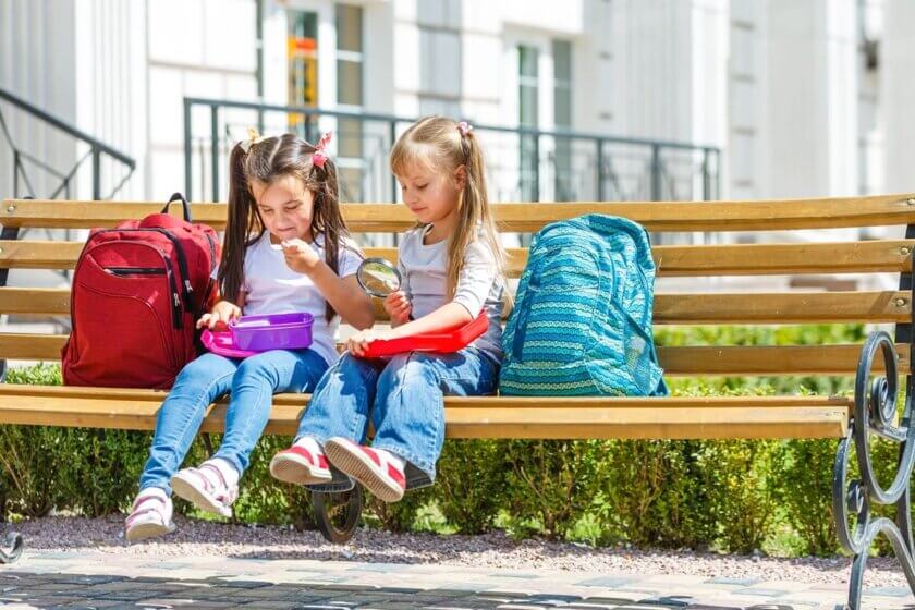 Zwei Grundschulkinder sitzen auf einer Schulbank und öffnen gemeinsam ihre Brotdosen in der Pause