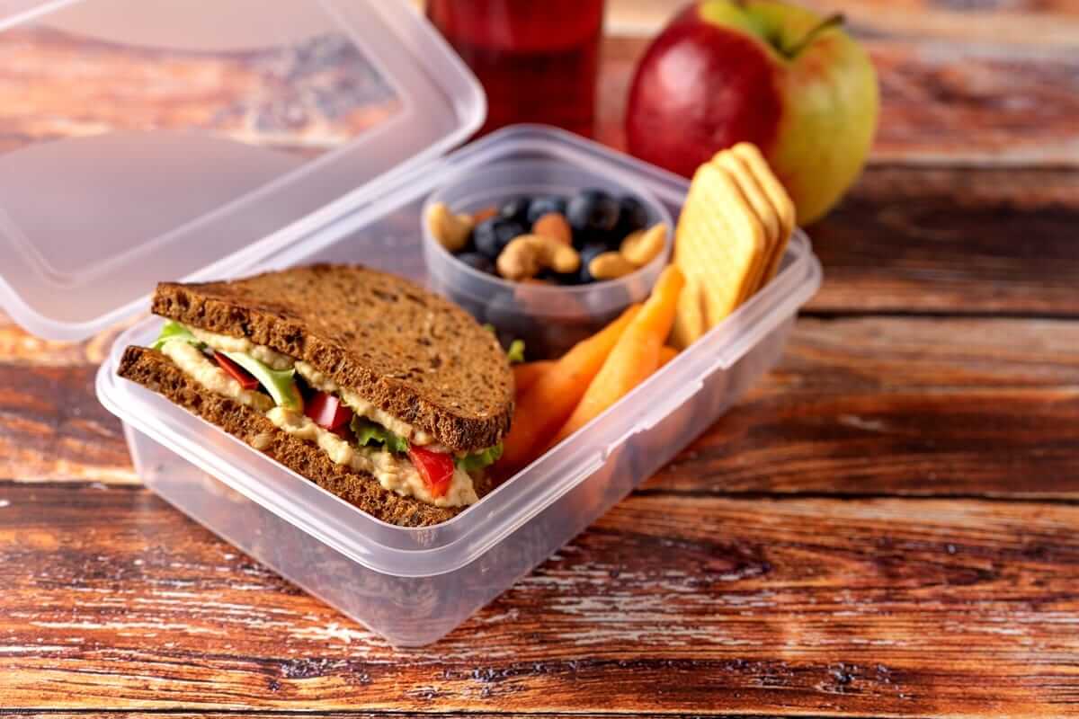Durchsichtige Brotdose mit belegtem Vollkornbrot, Möhren, Nüssen, Äpfeln daneben auf Holztisch