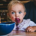 Beikost / Kleinkind sitzt vor einer blauen Schüssel mit brei-beschmiertem Gesicht und schaut direkt in die Kamera