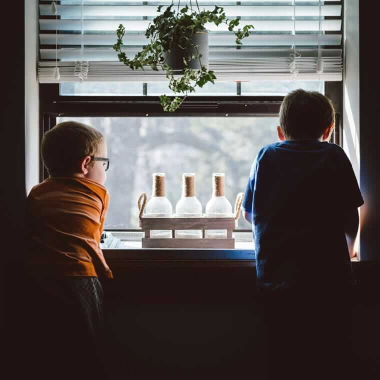 Sommerferien mit Kindern zu Hause: Ideen, Struktur und weniger Stress Langeweile / Zwei Jungen stehen nebeneinander am Fenster und schauen nach draußen – Kinder zuhause an einem Sommertag