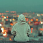 Sorgen, Eltern, Kinder / Teenager in Kapuzenpulli sitzt allein auf einer Mauer und blickt über eine beleuchtete Stadt – Symbol für kindliche Selbstständigkeit und die große Welt, die Eltern oft als bedrohlicher wahrnehmen als sie ist.