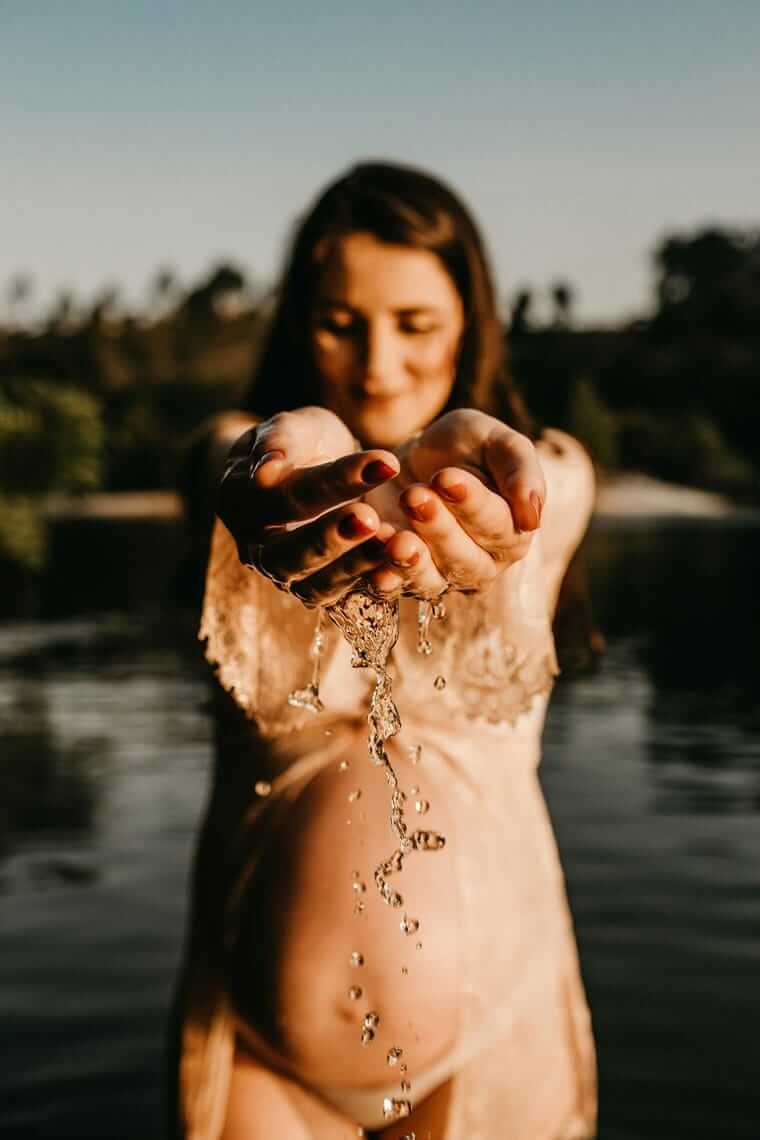 Schwanger bei Hitze / Wasser läuft durch die Hände einer schwangeren Frau im Gegenlicht, Fluss im Hintergrund