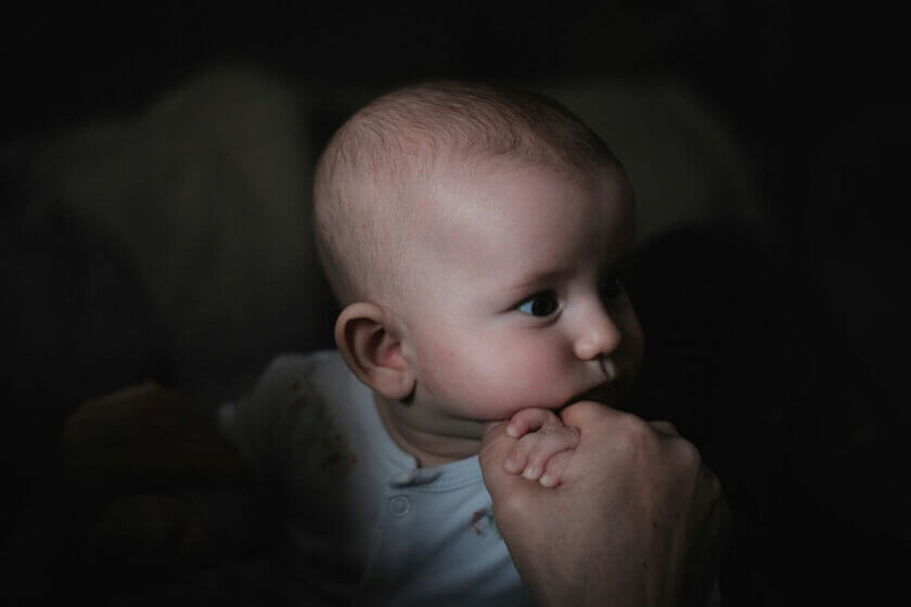 Baby schläft nicht ein trotz Müdigkeit / Säugling liegt im Dunkeln wach, schaut zur Seite, eine Erwachsenenhand hält ihn sanft