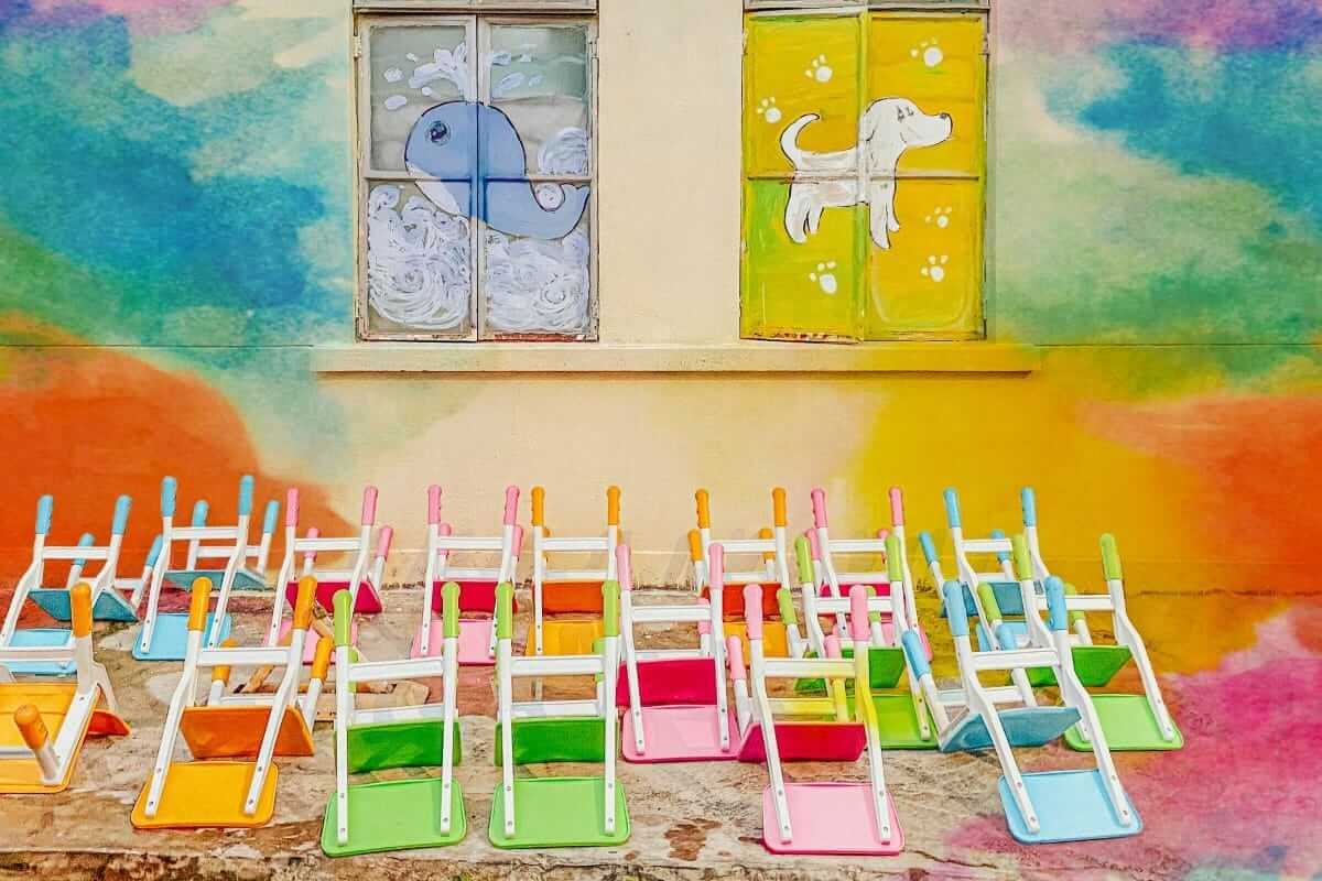 Wie findet man die perfekte Kita für sein Kind? Kita / Bunte Kinderstühle aufgereiht vor einer farbenfrohen Hausfassade mit gemalten Tiermotiven auf den Fenstern einer Kindertagesstätte