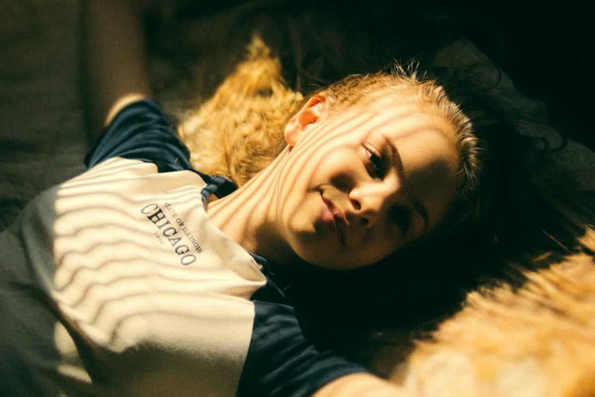 Pubertät bei Mädchen: Was passiert – und wie du begleitest Junges Mädchen liegt auf dem Boden, Lichtstreifen fallen über ihr Gesicht, entspannter Blick nach oben – Chicago-Shirt, warme Farbstimmung