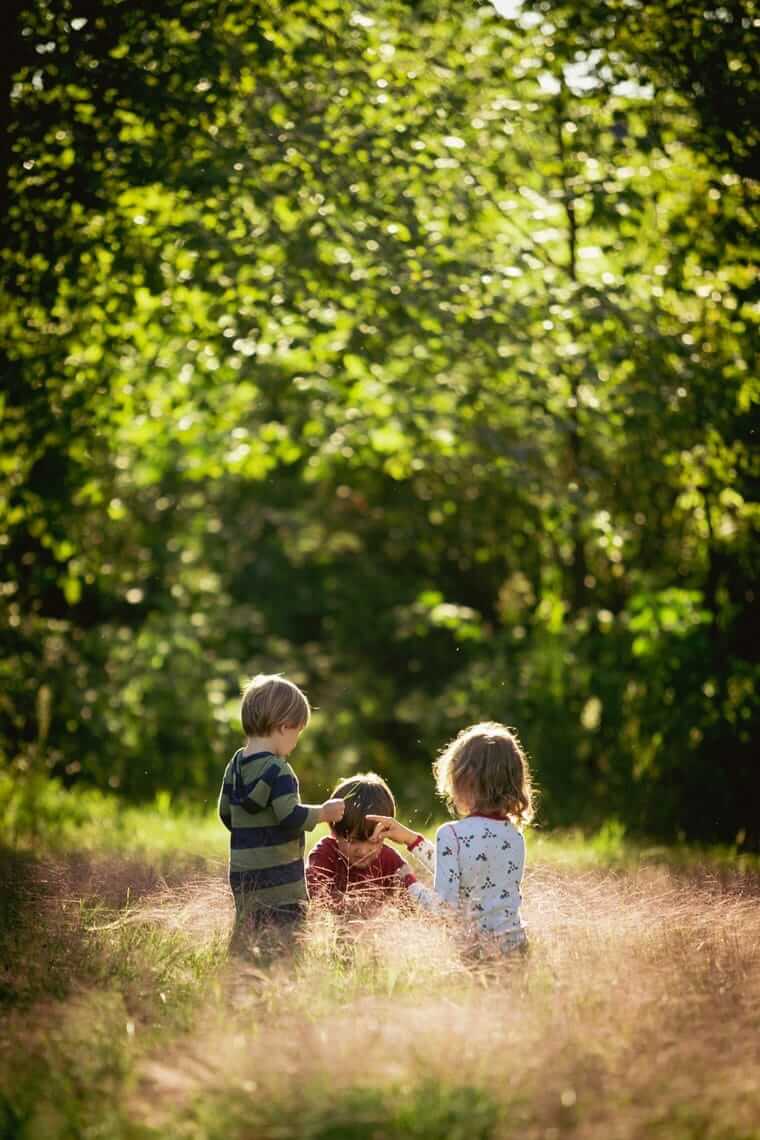 Sommerferien ohne Programm / Drei Kleinkinder sitzen von hinten auf einem Wiesenpfad im Gegenlicht eines Sommerabends und spielen miteinander.
