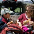 Snacks für unterwegs / Mutter und Kind sitzen auf einer Bank und essen Brötchen auf einem Ausflug in den Bergen – Rucksäcke neben sich