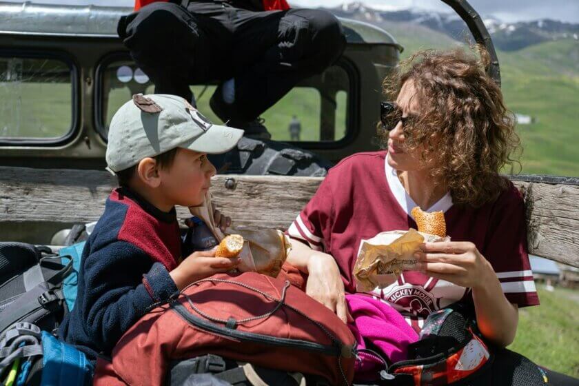 Snacks für unterwegs / Mutter und Kind sitzen auf einer Bank und essen Brötchen auf einem Ausflug in den Bergen – Rucksäcke neben sich