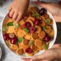 Erwachsene und Kinderhände an einer Schale mit Mini-Pancakes und frischen Kirschen – Snack für die ganze Familie.