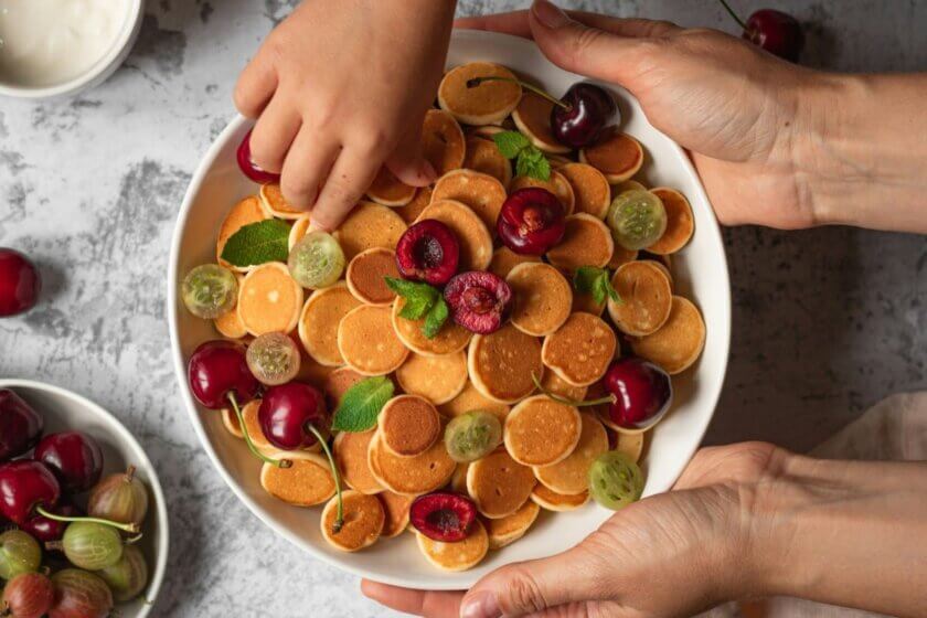 Erwachsene und Kinderhände an einer Schale mit Mini-Pancakes und frischen Kirschen – Snack für die ganze Familie.