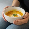 Zwei Hände halten eine Schale mit orangefarbener Kürbissuppe – Suppe als wärmendes Familienessen im Herbst
