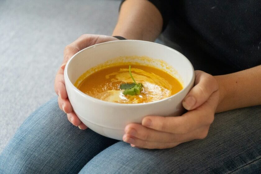 Zwei Hände halten eine Schale mit orangefarbener Kürbissuppe – Suppe als wärmendes Familienessen im Herbst