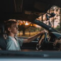 Junger Teenager sitzt konzentriert am Steuer eines Autos, Blick auf die Straße gerichtet, warmes Gegenlicht im Hintergrund.