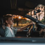Junger Teenager sitzt konzentriert am Steuer eines Autos, Blick auf die Straße gerichtet, warmes Gegenlicht im Hintergrund.