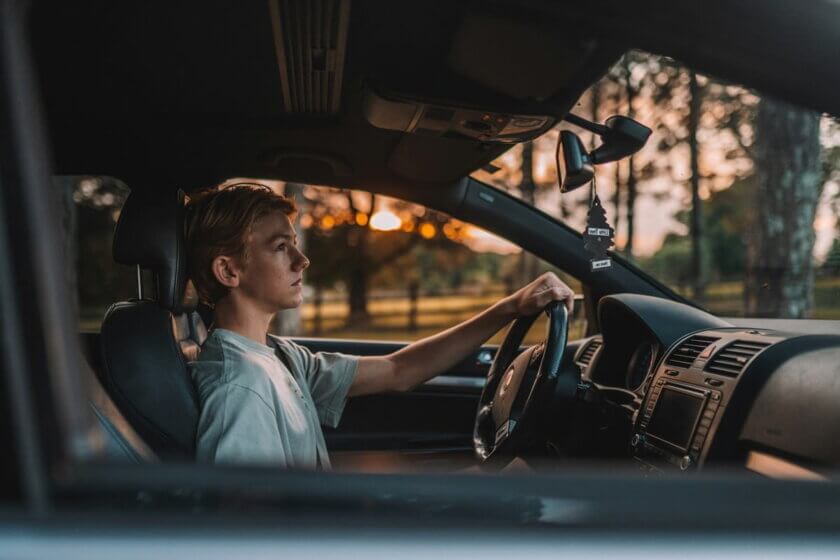 Junger Teenager sitzt konzentriert am Steuer eines Autos, Blick auf die Straße gerichtet, warmes Gegenlicht im Hintergrund.