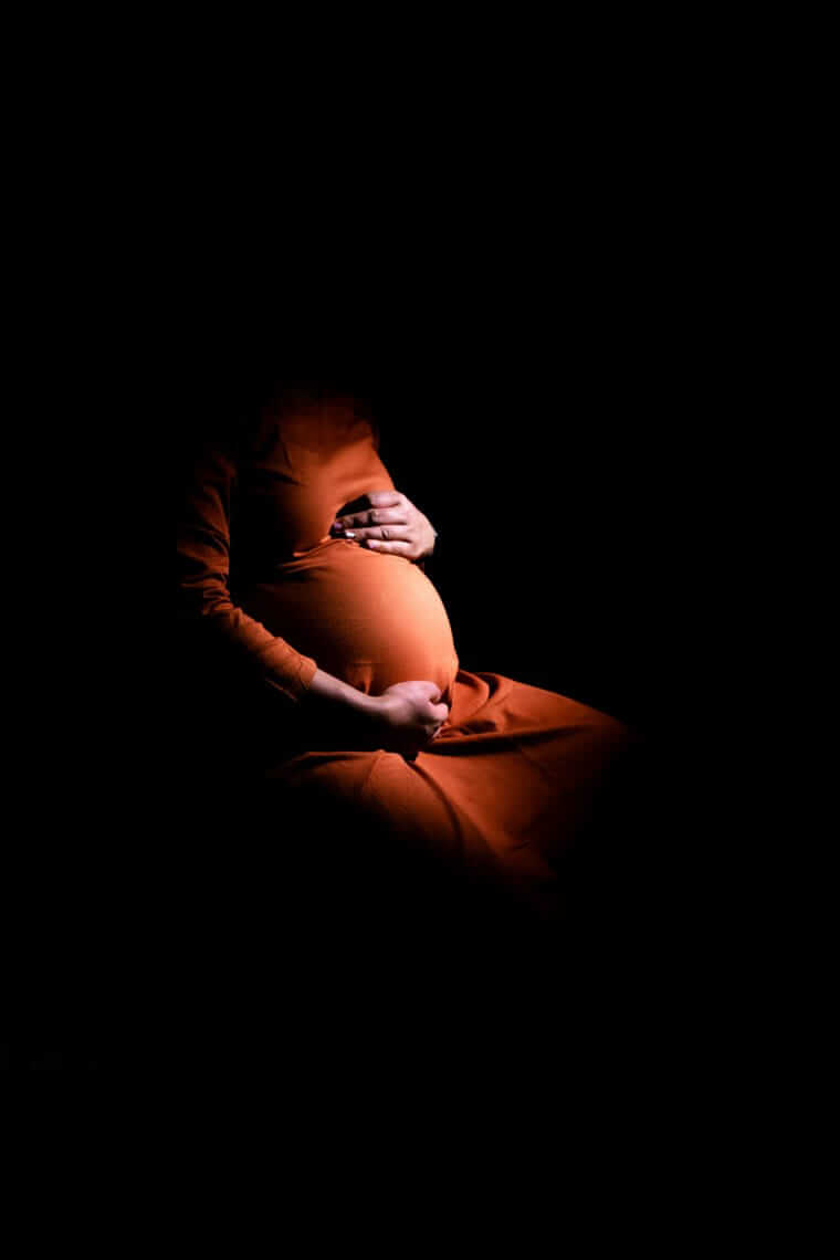 Schwangerschaftsdepression / Schwangere Frau im rostfarbenen Kleid hält ihren Bauch mit beiden Händen, vor dunklem Hintergrund, kein Gesicht sichtbar.