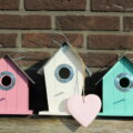 Nesting-Party / Drei bunte Vogelhäuschen in Rosa, Weiß und Türkis an einer Backsteinwand, in der Mitte hängt ein Herz aus Holz.
